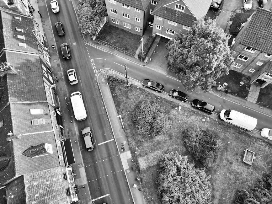 An aerial view of a residential street with a small grassy park area, featuring several parked cars along the curving road. The park contains a few trees and bushes, with pathways and a small square p