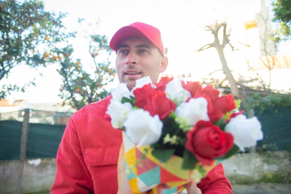A delivery person wearing a red cap and matching red jacket is holding a vibrant floral bouquet wrapped in patterned paper, featuring red and white roses with green foliage. The bouquet appears fresh,