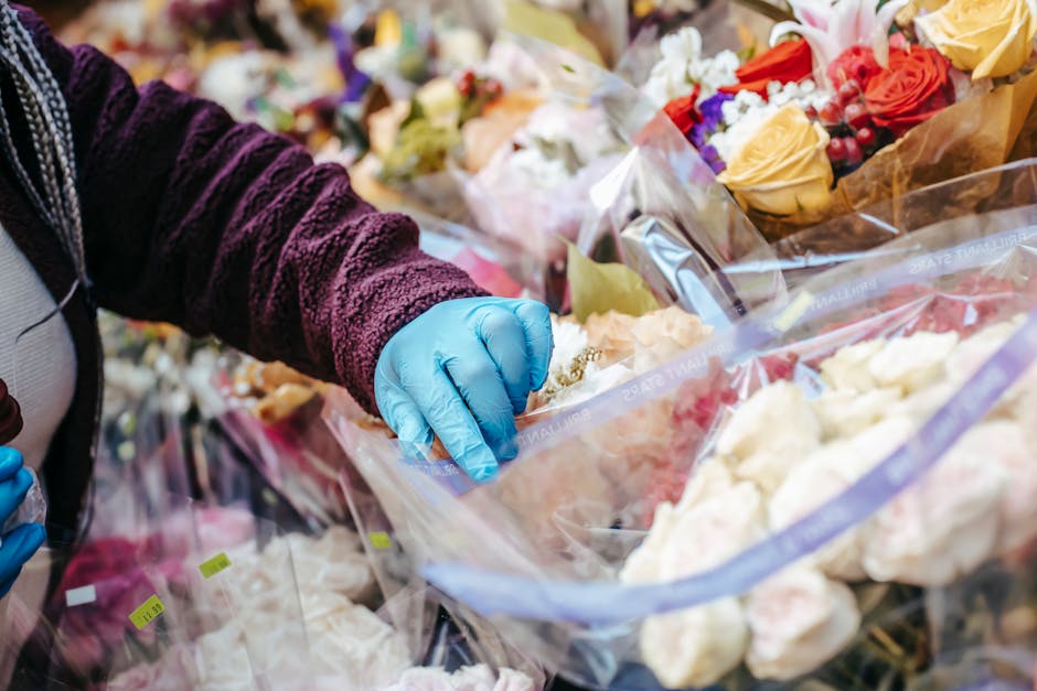 A person wearing a purple velvety jacket and blue disposable gloves arranges a bouquet of fresh flowers in clear plastic wrapping at a flower stall. The bouquet features peach roses, white roses, and