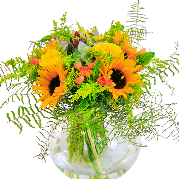 The first glance at this spirited bouquet from Flowers Purfleet captures an undeniable sense of radiant joy, as if a piece of the summer sun has found its way indoors. Dominating the display, vivid sunflowers unfurl their sunshine-yellow petals in striking contrast to dark, textural centres-the arrangement echoing scenes of wildflowers blooming near the Thames Path. Nearby, garden roses in creamy yellow spirals add a luxurious touch, their tightly layered petals reminiscent of traditional English rose gardens once enjoyed along London Road. Punctuating the design are clusters of bold red Hypericum berries, their glossiness enlivening the greenery and providing a cheerful, contemporary twist. Cascading ferns and delicate yellow goldenrod give the bouquet a wild, airy dynamism, reinforced by slender green spikes and the cool, understated presence of eucalyptus. Housed in a clear, spherical vase, the stems crisscross in water, their lines magnified and elongated by the glass-a modern statement grounded in Purfleet's fresh, riverside vibe. The scent is soft and inviting: roses mingling with herbal greenery, evoking a sense of open, sunlit fields. Whether intended for a joyful milestone or simply to bring a smile to someone in Purfleet, this arrangement is a glowing tribute to the natural beauty and local artistry that flourish along the Thames.